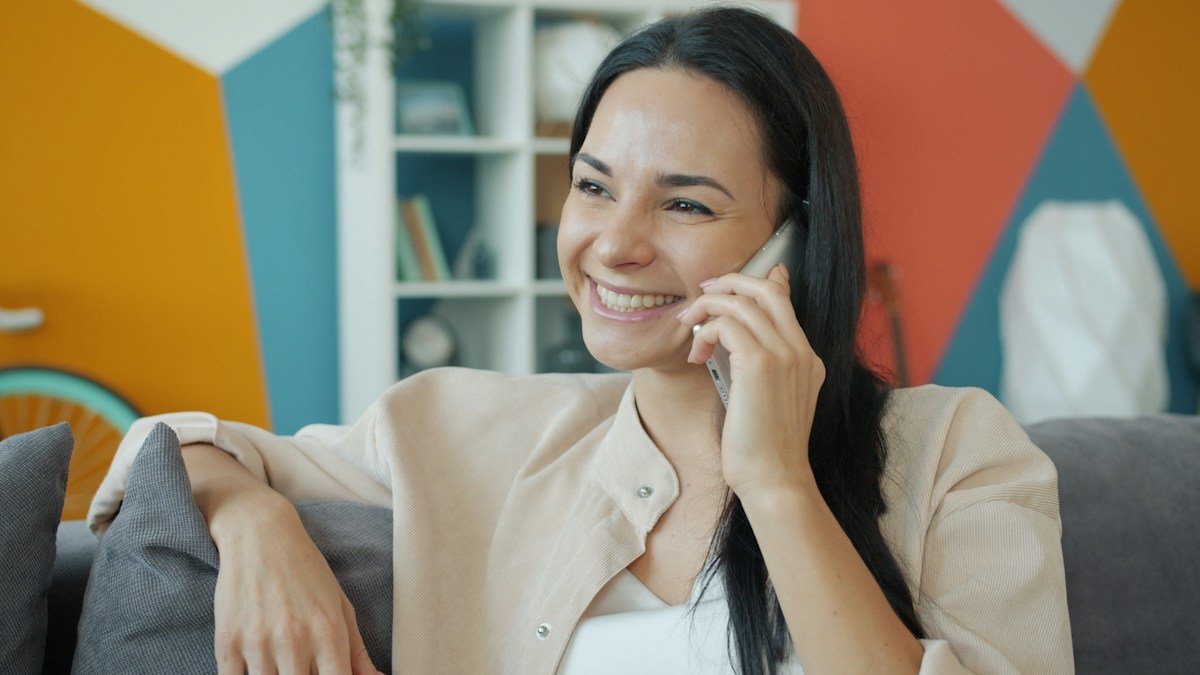 Frau laechelt waehrend eines Telefonats mit dem Handy am Ohr zu Hause